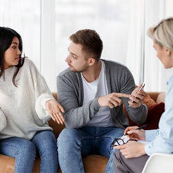 A couple appear to argue while someone holds and points at a phone in front of a third person. Learn more about the support a couples therapist in California can offer by searching "couples retreat in California" or "infidelity therapist near me"