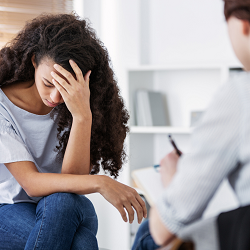 A woman holds her head while a person with a clipboard taking notes. Learn more about trauma therapy in Roseville, CA and the support we can offer for PTSD in Fair Oaks, CA. Contact a therapist for teens in Roseville, CA to learn more today.